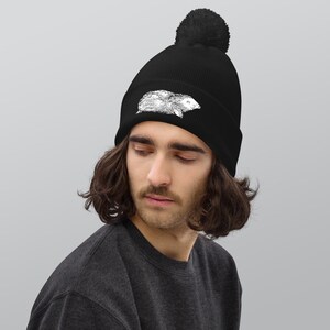 May include: Black beanie with a white illustration of a rodent. The beanie has a ribbed knit design and a black pom-pom on top. The hat is worn by a person with long, wavy hair.