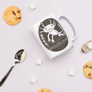 Puede incluir: Taza de cer&aacute;mica blanca con un dise&ntilde;o de ajolote en blanco y negro dentro de un &oacute;valo gris oscuro. El texto "AXOLOTL MOM" est&aacute; debajo. La taza est&aacute; rodeada de galletas, terrones de az&uacute;car y una cuchara.