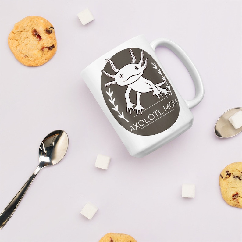 Puede incluir: Taza de cer&aacute;mica blanca con un dise&ntilde;o de ajolote en blanco y negro dentro de un &oacute;valo gris oscuro. El texto "AXOLOTL MOM" est&aacute; debajo. La taza est&aacute; rodeada de galletas, terrones de az&uacute;car y una cuchara.