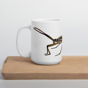 Peut inclure: Mug en céramique blanche avec une illustration détaillée d'un insecte brun et jaune. L'insecte est représenté de profil, avec de longues antennes et des pattes fines. La tasse a une grande anse et une finition brillante.