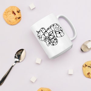 K&ouml;nnte beinhalten: Wei&szlig;e Keramiktasse mit einer schwarz-wei&szlig;en Illustration einer Schlange. Die Tasse hat einen gro&szlig;en Henkel und ist von Keksen, Zuckerw&uuml;rfeln und einem L&ouml;ffel umgeben. Ein tolles Geschenk f&uuml;r Reptilienliebhaber.