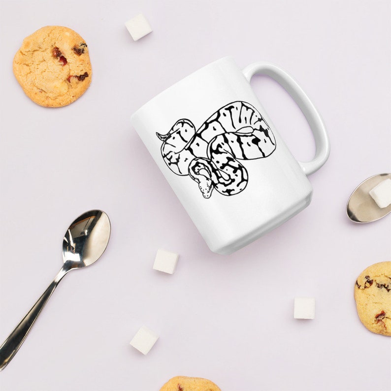K&ouml;nnte beinhalten: Wei&szlig;e Keramiktasse mit einer schwarz-wei&szlig;en Illustration einer Schlange. Die Tasse hat einen gro&szlig;en Henkel und ist von Keksen, Zuckerw&uuml;rfeln und einem L&ouml;ffel umgeben. Ein tolles Geschenk f&uuml;r Reptilienliebhaber.