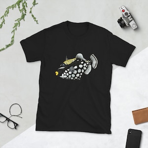May include: Black t-shirt featuring a detailed illustration of a clown triggerfish. The fish has a black body with white spots, yellow accents, and a yellow mouth. The design is centered on the shirt.