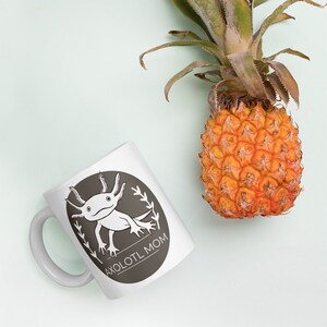 Puede incluir: Taza de cer&aacute;mica blanca con un dise&ntilde;o ovalado gris que presenta un ajolote de dibujos animados y el texto "AXOLOTL MOM". Una pi&ntilde;a madura con hojas verdes se encuentra a la derecha de la taza sobre un fondo azul claro.