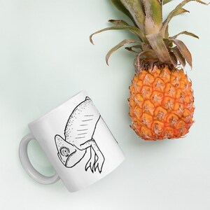 Puede incluir: Taza de cerámica blanca con un dibujo lineal en blanco y negro de un camaleón. Una piña madura con hojas verdes está junto a la taza sobre un fondo azul claro. La taza tiene un asa curva.