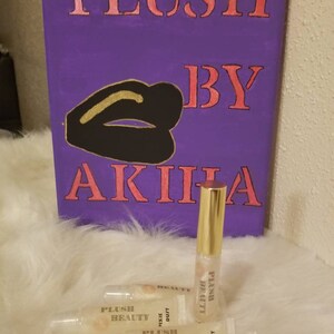 Peut inclure: Une toile violette avec les mots "PLUSH BY AKIHA" en rose et un dessin de l&egrave;vres noir et or. Plusieurs tubes de brillant &agrave; l&egrave;vres transparents avec des bouchons dor&eacute;s et le texte "PLUSH BEAUTY PIXIE DUST" sont devant la toile.