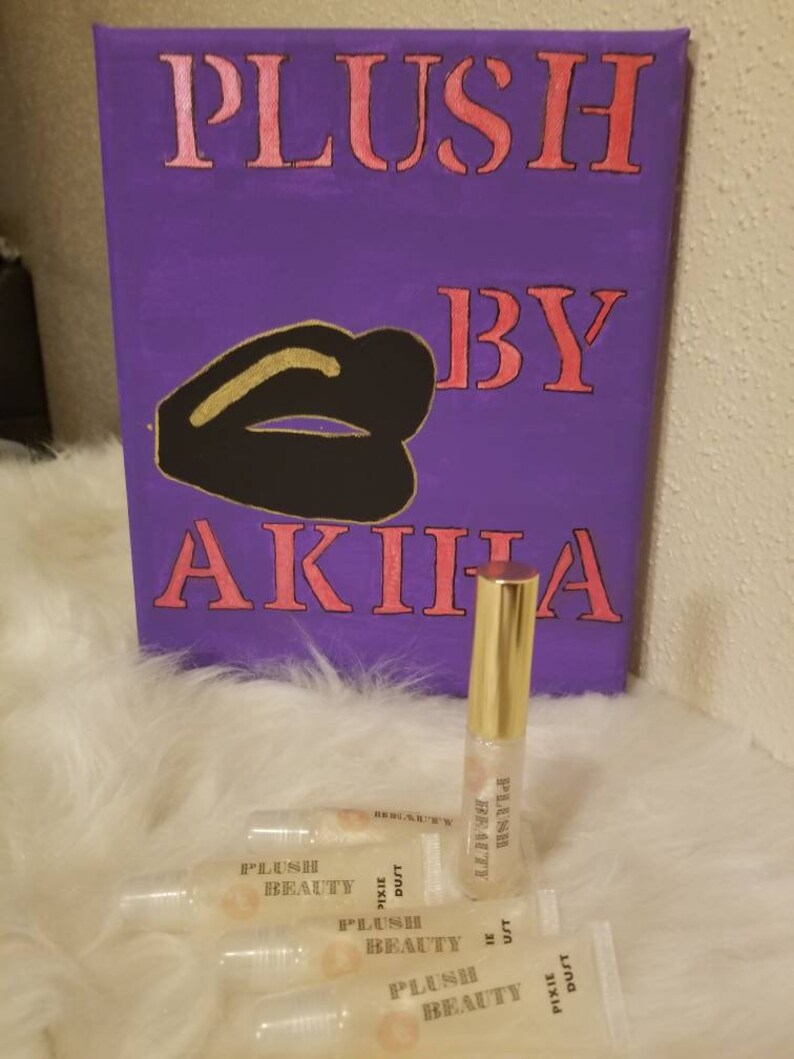 Peut inclure: Une toile violette avec les mots "PLUSH BY AKIHA" en rose et un dessin de l&egrave;vres noir et or. Plusieurs tubes de brillant &agrave; l&egrave;vres transparents avec des bouchons dor&eacute;s et le texte "PLUSH BEAUTY PIXIE DUST" sont devant la toile.