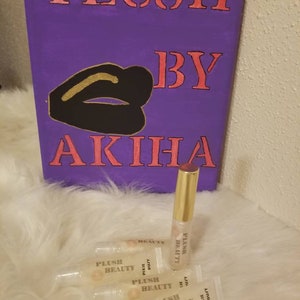 May include: A purple canvas with the words "PLUSH BY AKIHA" in pink lettering, and a black lip design with gold accents. Several lip gloss tubes with the text "PLUSH BEAUTY" and "PIXIE DUST" are in front of the canvas.