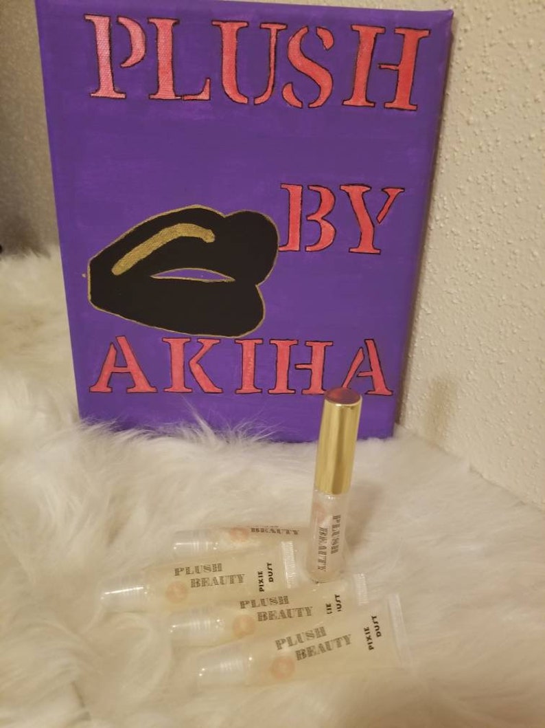 Peut inclure: Une toile violette avec les mots "PLUSH BY AKIHA" en lettres roses, et un dessin de l&egrave;vres noires avec des accents dor&eacute;s. Plusieurs tubes de gloss avec le texte "PLUSH BEAUTY" et "PIXIE DUST" sont devant la toile.
