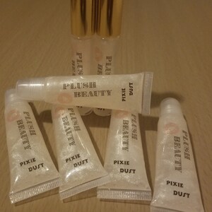 Peut inclure: Tubes de brillant &agrave; l&egrave;vres transparents avec des bouchons dor&eacute;s et le texte "PLUSH BEAUTY PIXIE DUST". Les tubes sont dispos&eacute;s sur une surface en bois. Le brillant &agrave; l&egrave;vres a un aspect scintillant et paillet&eacute;.