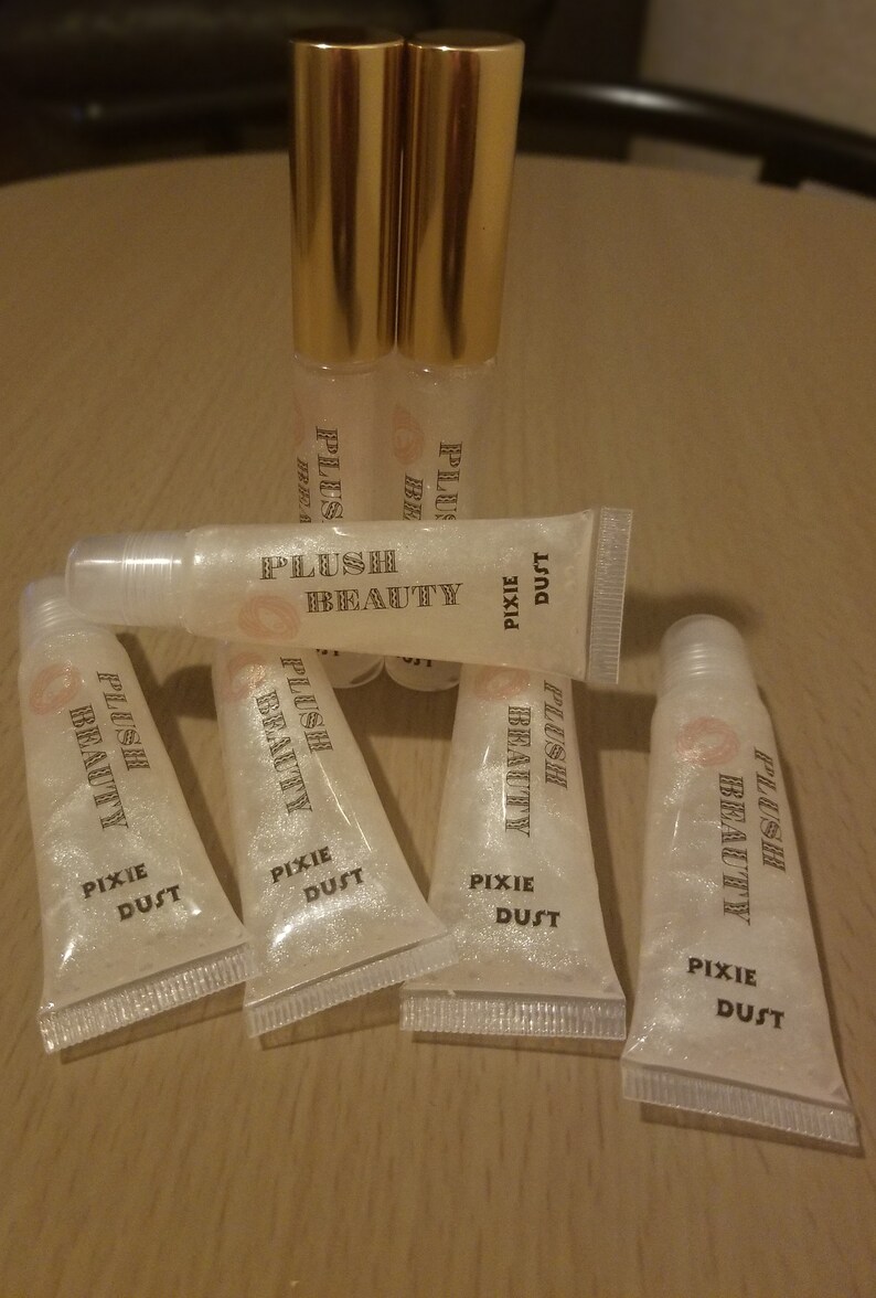 Peut inclure: Tubes de brillant &agrave; l&egrave;vres transparents avec des bouchons dor&eacute;s et le texte "PLUSH BEAUTY PIXIE DUST". Les tubes sont dispos&eacute;s sur une surface en bois. Le brillant &agrave; l&egrave;vres a un aspect scintillant et paillet&eacute;.