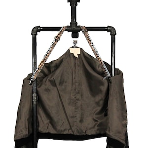 Könnte beinhalten: Ein schwarzer Metall-Kleiderständer mit einem Leopardenmuster-Band, an dem eine braune Jacke mit schwarzen Ärmeln hängt.