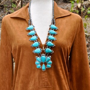 May include: A brown suede-like dress with a turquoise squash blossom necklace and matching belt. The necklace and belt feature silver settings and teardrop-shaped turquoise stones. The dress has a collared neckline and long sleeves.