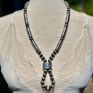 May include: A silver necklace featuring a series of small, round beads and elongated, cylindrical beads with etched designs. The necklace has a central pendant with a geometric pattern and a teardrop shape.