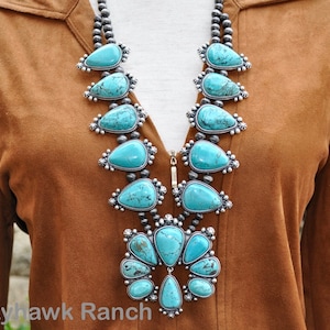 May include: A turquoise squash blossom necklace with silver accents. The necklace features large, teardrop-shaped turquoise stones set in ornate silver settings, strung on a strand of small, round beads. The necklace is displayed against a brown background.