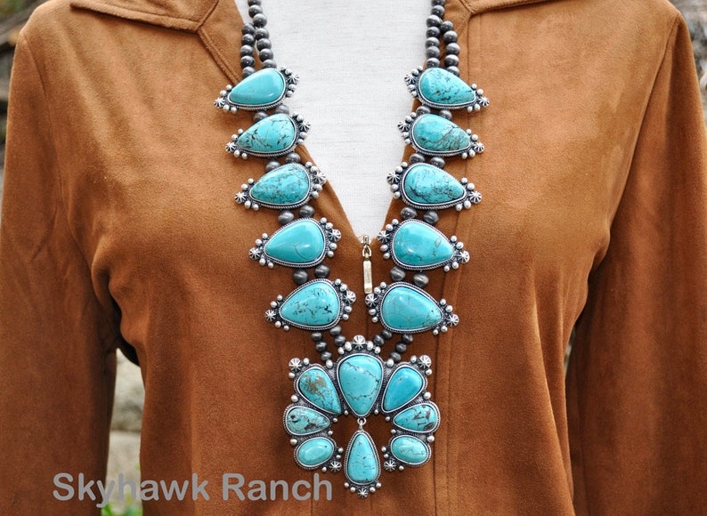 May include: A turquoise squash blossom necklace with silver accents. The necklace features large, teardrop-shaped turquoise stones set in ornate silver settings, strung on a strand of small, round beads. The necklace is displayed against a brown background.