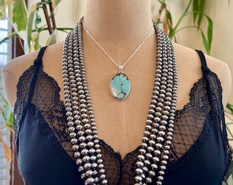 Puede incluir: Un collar de múltiples hilos con cuentas plateadas pulidas y un colgante ovalado central con una piedra turquesa azul claro con patrones de matriz marrón, suspendido de una delicada cadena plateada sobre una blusa negra con encaje.