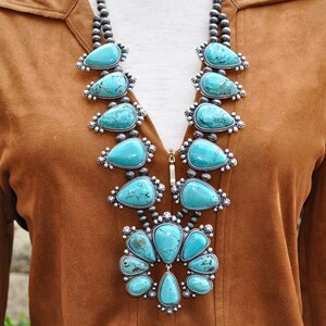 May include: A turquoise squash blossom necklace with silver accents. The necklace features large, teardrop-shaped turquoise stones set in ornate silver settings, connected by strands of small silver beads. The necklace is displayed against a brown background.