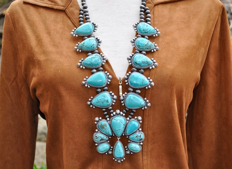 May include: A turquoise squash blossom necklace with silver accents. The necklace features large, teardrop-shaped turquoise stones set in ornate silver settings, connected by strands of small silver beads. The necklace is displayed against a brown background.