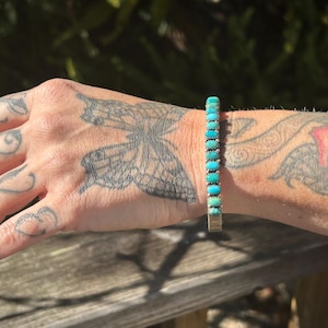 May include: A turquoise and silver bracelet with a row of turquoise stones. The bracelet is on a person's wrist.