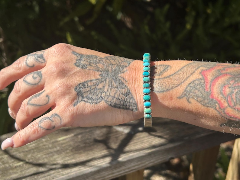 May include: A turquoise and silver bracelet with a row of turquoise stones. The bracelet is on a person's wrist.