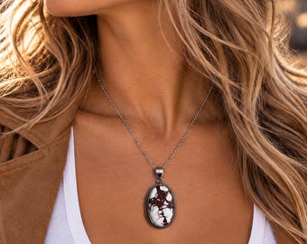May include: A silver necklace featuring an oval pendant with a white and dark brown marbled stone set in a textured metal frame. The pendant hangs from a delicate silver chain, resting against a white tank top and a brown suede jacket.