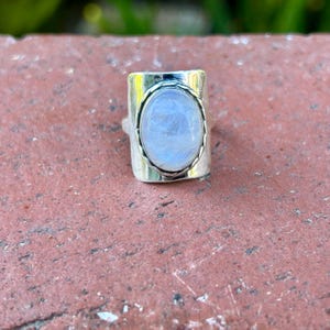 Puede incluir: Un anillo de plata con una gran piedra de luna ovalada engastada en un bisel rectangular. El anillo tiene una banda texturizada.