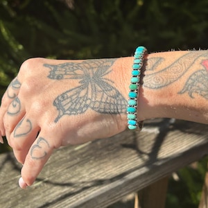 May include: A silver bracelet with turquoise stones. The stones are oval shaped and graduated in size, with the largest stones in the center of the bracelet. The bracelet is on a person's wrist.