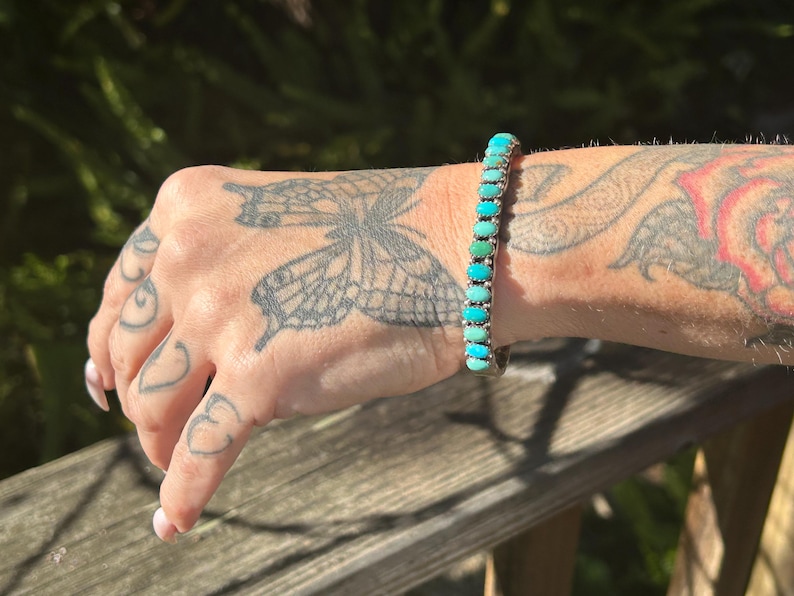 May include: A silver bracelet with turquoise stones. The stones are oval shaped and graduated in size, with the largest stones in the center of the bracelet. The bracelet is on a person's wrist.