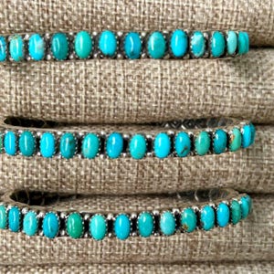 May include: Three turquoise and silver bracelets stacked on top of each other. The bracelets are made of silver with turquoise stones set in the metal. The bracelets are on a brown fabric background.