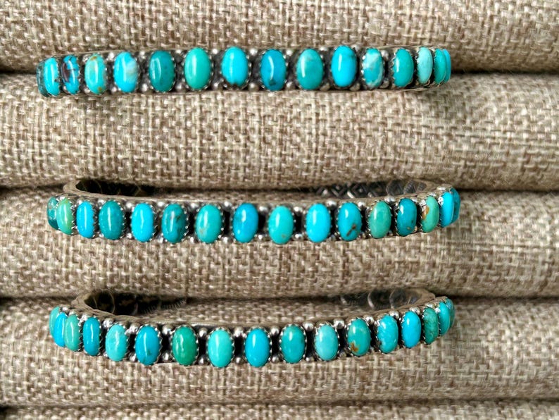 May include: Three turquoise and silver bracelets stacked on top of each other. The bracelets are made of silver with turquoise stones set in the metal. The bracelets are on a brown fabric background.