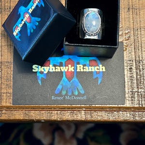 Puede incluir: Un anillo de plata con una gran piedra de luna ovalada engastada en un engaste de bisel. El anillo est&aacute; en una caja negra con un logotipo que dice "Skyhawk Ranch" y "Renee' McDonnell".