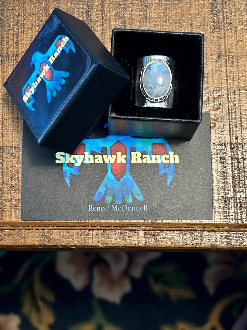 Puede incluir: Un anillo de plata con una gran piedra de luna ovalada engastada en un engaste de bisel. El anillo est&aacute; en una caja negra con un logotipo que dice "Skyhawk Ranch" y "Renee' McDonnell".