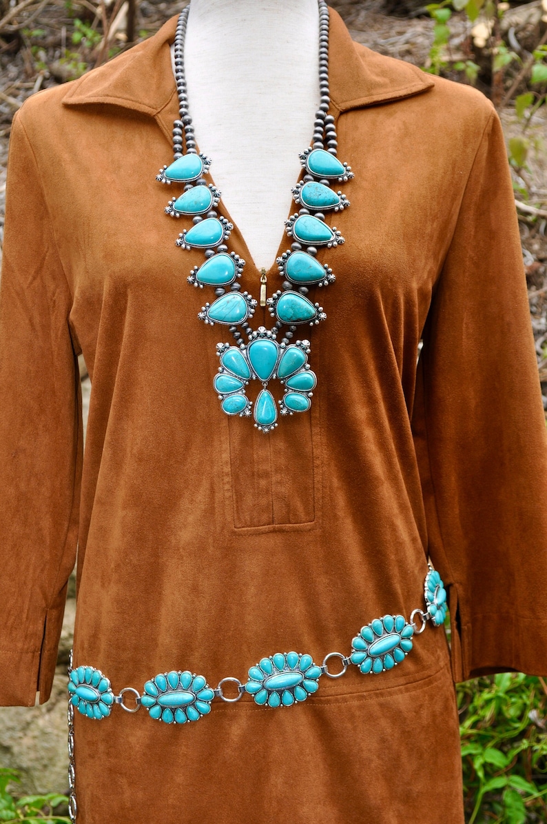 May include: A brown suede-like dress with a turquoise squash blossom necklace and matching belt. The necklace and belt feature silver settings and teardrop-shaped turquoise stones. The dress has a collared neckline and long sleeves.