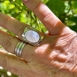Puede incluir: Un anillo de plata con una gran piedra de luna ovalada engastada en un bisel rectangular. El anillo est&aacute; en un dedo con una banda de plata en el dedo de al lado.