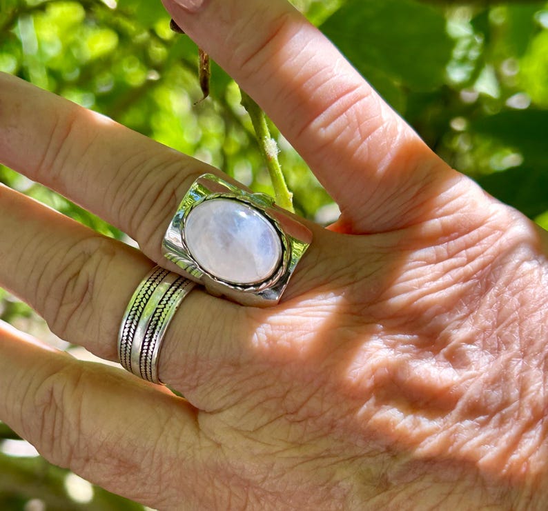 Puede incluir: Un anillo de plata con una gran piedra de luna ovalada engastada en un bisel rectangular. El anillo est&aacute; en un dedo con una banda de plata en el dedo de al lado.