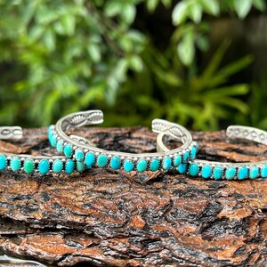 May include: Three silver bracelets with turquoise stones. The bracelets are stacked on top of each other and are on a brown wooden surface.