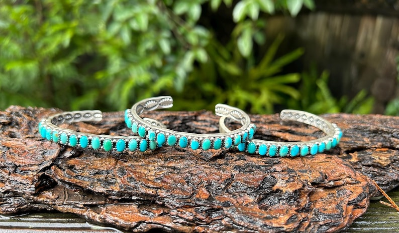 May include: Three silver bracelets with turquoise stones. The bracelets are stacked on top of each other and are on a brown wooden surface.