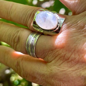 Puede incluir: Un anillo de plata con una gran piedra de luna ovalada. El anillo tiene una banda texturizada y se lleva en un dedo.