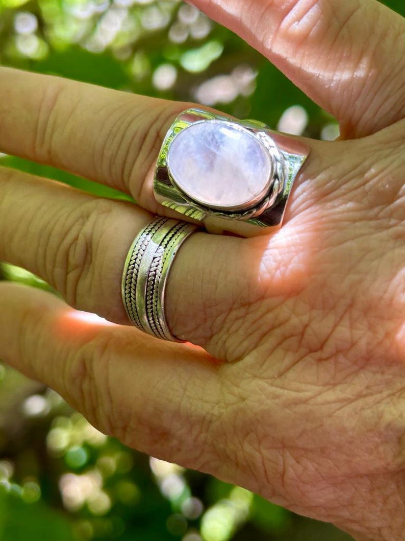 Puede incluir: Un anillo de plata con una gran piedra de luna ovalada. El anillo tiene una banda texturizada y se lleva en un dedo.