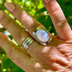 Puede incluir: Un anillo de plata con una gran piedra de luna ovalada engastada en un engaste de bisel. El anillo se lleva en un dedo con un anillo de banda de plata en el dedo de al lado.