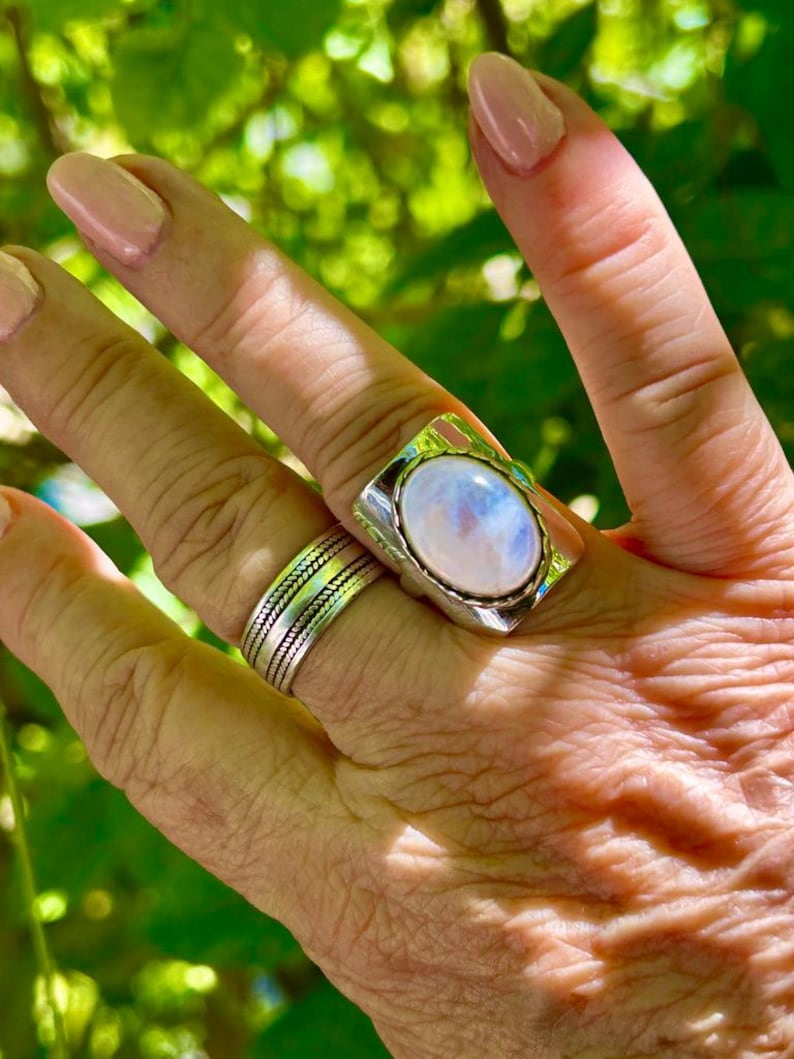 Puede incluir: Un anillo de plata con una gran piedra de luna ovalada engastada en un engaste de bisel. El anillo se lleva en un dedo con un anillo de banda de plata en el dedo de al lado.