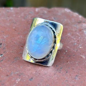Puede incluir: Un anillo de plata con una gran piedra de luna ovalada engastada en un bisel rectangular. El anillo tiene una banda texturizada.