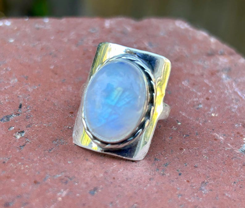 Puede incluir: Un anillo de plata con una gran piedra de luna ovalada engastada en un bisel rectangular. El anillo tiene una banda texturizada.