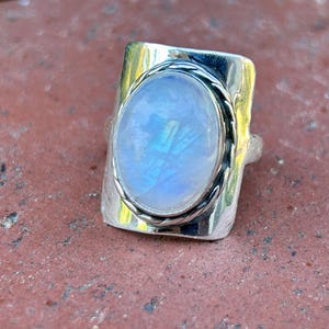 Puede incluir: Un anillo de plata con una gran piedra de luna ovalada engastada en un bisel texturizado. El anillo tiene una banda ancha con un dise&ntilde;o de cuerda.