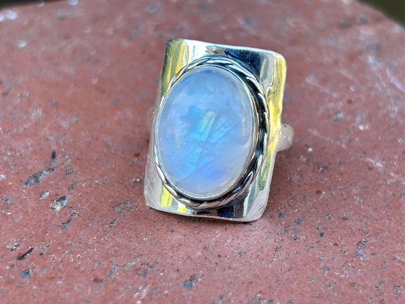 Puede incluir: Un anillo de plata con una gran piedra de luna ovalada engastada en un bisel texturizado. El anillo tiene una banda ancha con un dise&ntilde;o de cuerda.