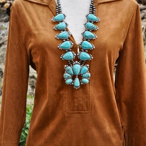 May include: A brown suede tunic with a collared neckline and long sleeves. A turquoise and silver squash blossom necklace is the focal point. The necklace features large, teardrop-shaped turquoise stones.