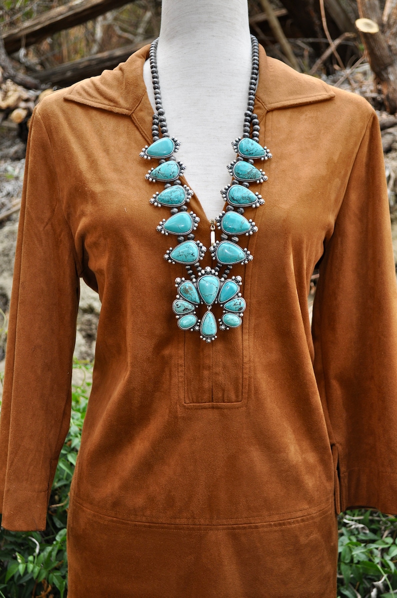 May include: A brown suede tunic with a collared neckline and long sleeves. A turquoise and silver squash blossom necklace is the focal point. The necklace features large, teardrop-shaped turquoise stones.