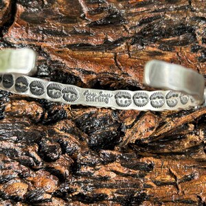 May include: A silver bracelet with a turquoise stone and a stamped design. The bracelet is stamped "Larry Goins Sterling".