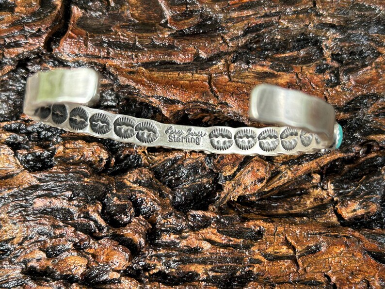May include: A silver bracelet with a turquoise stone and a stamped design. The bracelet is stamped "Larry Goins Sterling".